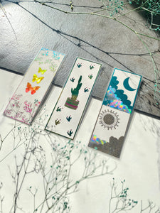 3 bookmarks. On the left bookmark with 3 yellow and orange butterflies and flowers. In the middle Green cactus bookmark. On the right bookmark with blue moon and gold sun