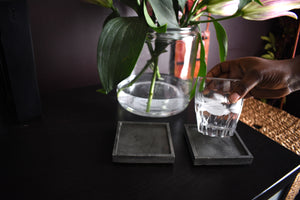 photo of 2 gray square coasters with plant in the background and a hand holding a glass of water over the coasters
